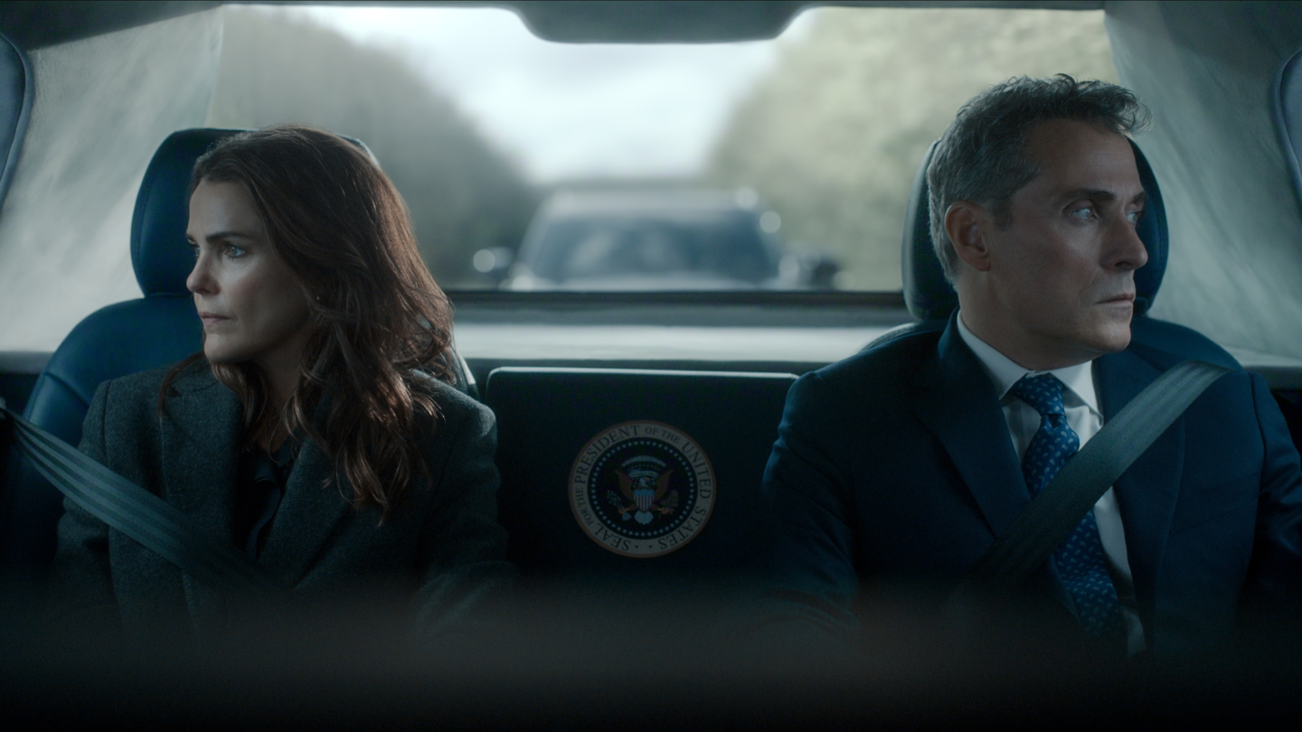 "Two serious people in formal attire sit in the back of a car with a presidential seal, looking out the windows; the environment is tense, with another vehicle visible behind them through the rear window."